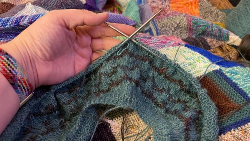 A few inches of knitted sweater yoke in teal and varigated yarn, being held over a knitted blanket